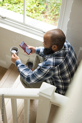 African American man sitting on stair landing by window holding red smartphone and mug, copy space