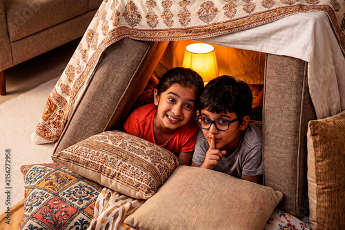 Cozy Blanket Fort Adventure: Two children share a secret moment of fun and imagination inside a makeshift blanket fort, illuminated by a soft lamp, surrounded by cushions.