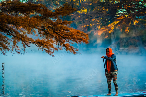 Woman paddleboards calm lake, mist curls around her