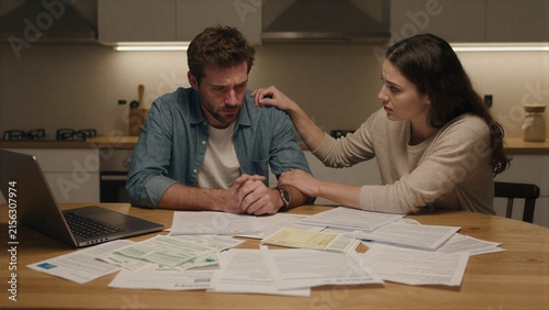 Couple sits at kitchen table, overwhelmed by bills and laptop.