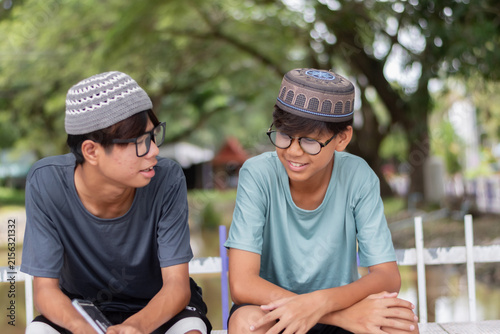 Two smiling Muslim teenage boys wearing traditional caps and talking together while sitting in a park with a blurred green background.