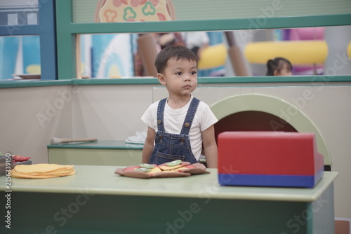 Obraz na plátně Cute toddler boy standing at play kitchen counter roleplaying in indoor playgrou
