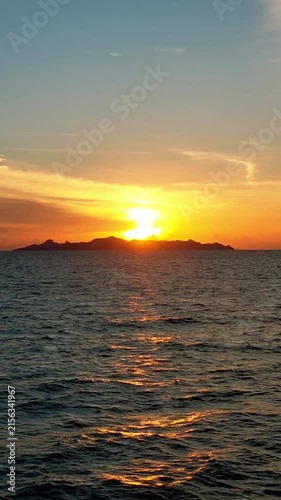 Beautiful vertical video of a golden sunset over the sea with an island silhouette on the horizon. The bright sun reflects on the rippling ocean waves.