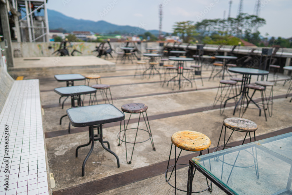 Fototapeta premium Industrial outdoor cafe setup featuring a round glass-top table surrounded by minimalist bar stools with wooden seats and black metal wire legs on a textured concrete pavement in bright daylight.