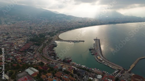 Alanya, Turkey. Aerial panoramic view of the historic harbor, Red Tower fortress, and Mediterranean coastline with dramatic sun rays piercing low clouds. Aerial View