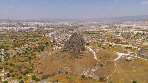 Goreme, Nevsehir, Turkey. A solitary volcanic tuff monolith rises above the arid Goreme Valley, an aerial panoramic view over eroded cliffs and scattered greenery under clear summer skies. Aerial Vie