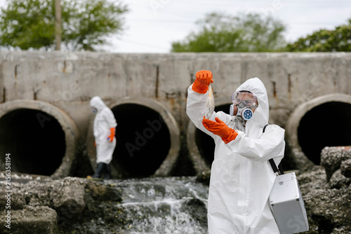Quadro su tela Scientists in hazmat suits collect water samples near concrete drainage pipes discharging polluted wastewater, testing contamination and environmental hazards at an outfall site