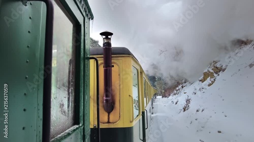Old steam train travels through a snowy mountain landscape, belching smoke and steam in scenic winter motion.