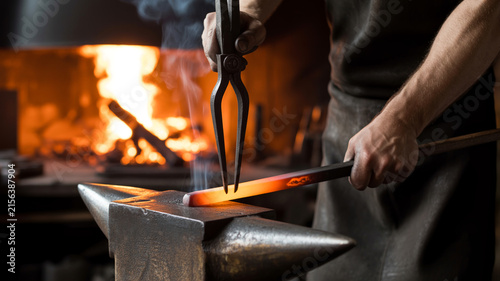 Blacksmith holding glowing red hot steel bar with tongs on anvil in traditional forge workshop stock photo
