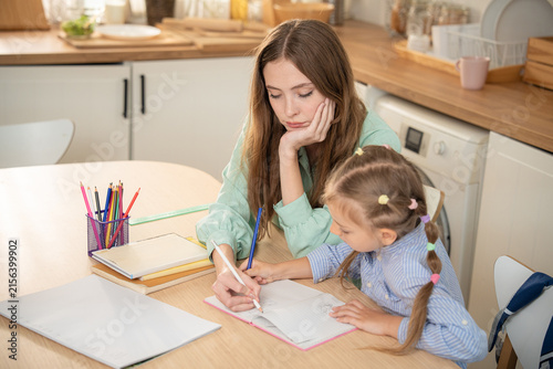 Young mother assisting small daughter with drawing and homework on table at home. Mother and little girl sitting together at kitchen table doing homeschool assignment with colored pencils and notebook