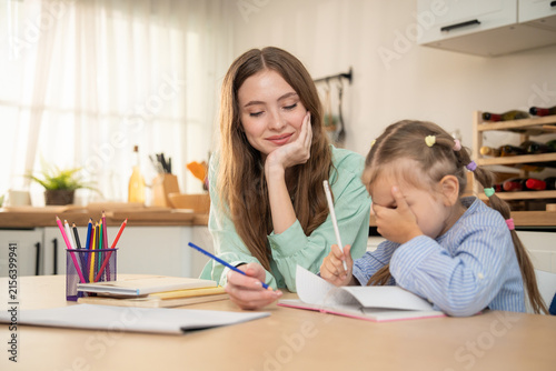 Mother gently watching her stressed daughter covering face while doing difficult homework. Mother sitting at kitchen table supporting daughter who feels frustrated and overwhelmed by home schooling.