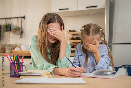 Stressed mother and daughter both covering face with hands while struggling with homework. Exhausted mother and little girl showing total frustration and fatigue during hard homeschooling lesson at ho