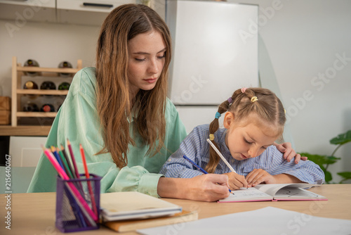 Little girl excitedly talking while writing homework with mother's supportive hand on her shoulder. Young girl enjoying homeschooling study time with her mother providing guidance and care at home.