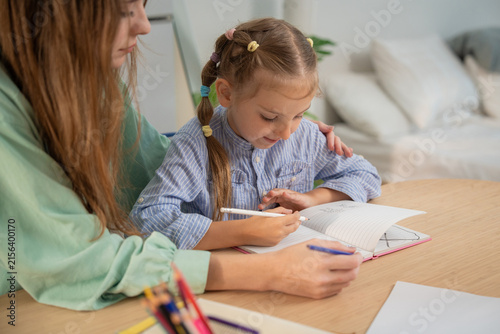Little girl excitedly talking while writing homework with mother's supportive hand on her shoulder. Young girl enjoying homeschooling study time with her mother providing guidance and care at home.