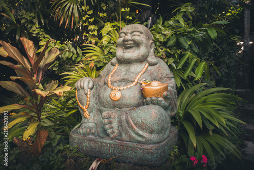 Laughing Buddha or Budai statue sitting in a lush tropical garden, representing happiness, good fortune, and prosperity in Asian culture and spirituality