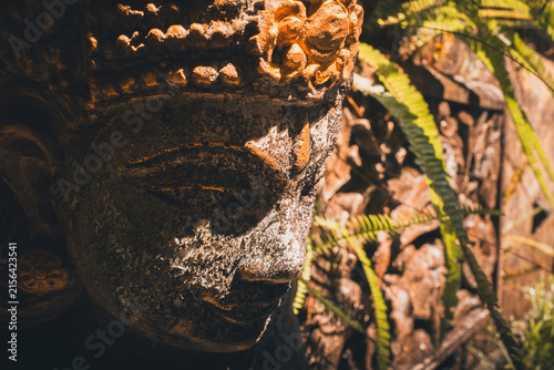 Weathered stone carving depicting a traditional Hindu deity, standing among lush tropical foliage in Bali, showing cultural heritage and ancient spiritual art