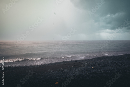 Moody stormy ocean scene with dark clouds bringing heavy rain, creating a dramatic atmosphere over the black volcanic beach and crashing waves