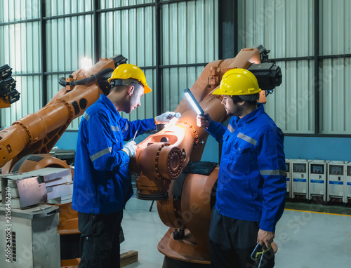 Two professional engineer blue safety uniform hardhat use inspection light bar inspect perform maintenance on heavy orange industrial robotic arm inside automated factory workspace.