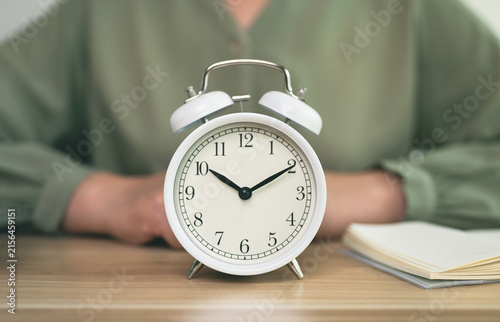 Closeup white alarm clock on desk with professional woman sitting on background. concept of representing office productivity, time management, deadline, punctuality, and work life balance.