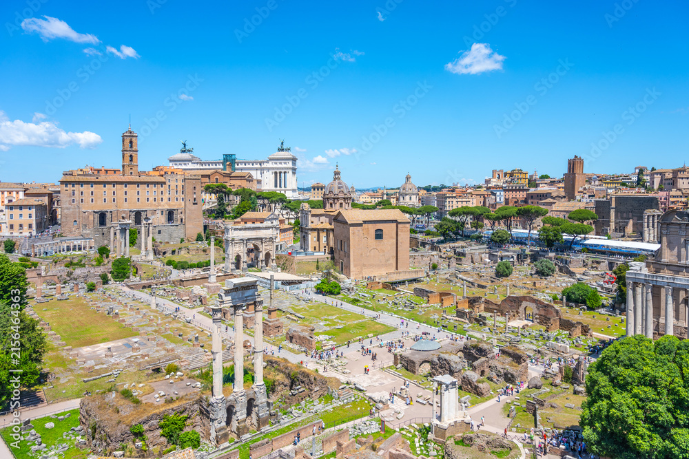 Fototapeta premium Tourists walk among ancient ruins at the Roman Forum in Rome Italy. The historic site shows remains of buildings and columns under a blue sky with few clouds.