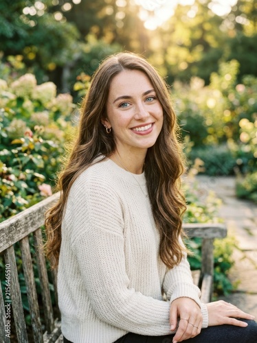 A young woman sits on a wooden bench in a rose garden. She is smiling and looking directly at the camera.