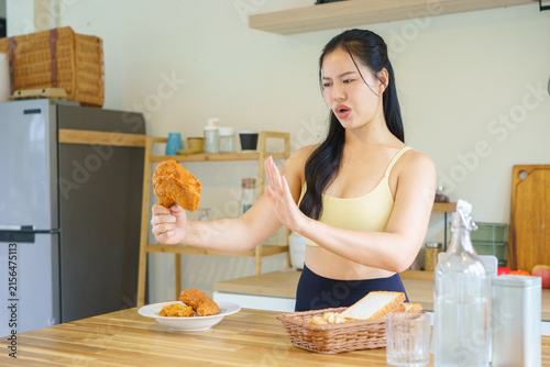 Obraz na plátně Fit Asian woman in yellow sports bra gestures refusal toward fried chicken in a modern kitchen, representing dieting, willpower, and healthy lifestyle choices