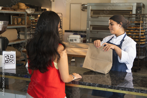 Bakery staff wearing chef coat handing paper bag to customer at counter, QR code, copy space