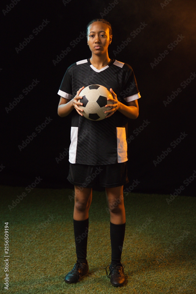 Fototapeta premium African adult woman standing on green turf studio-lit holding black-white soccer ball in black kit