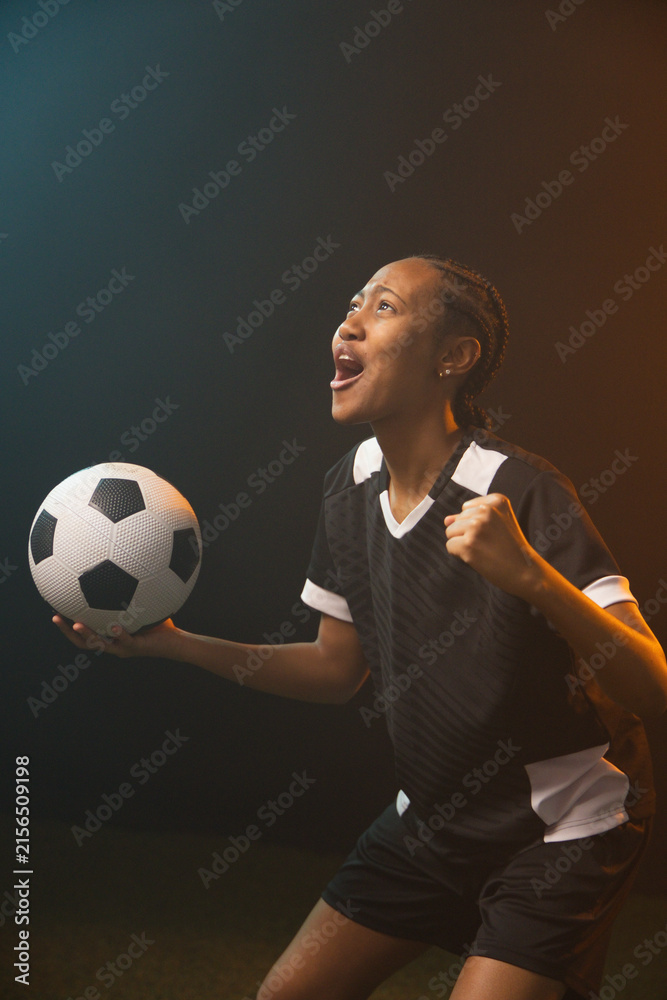 Fototapeta premium African American woman soccer player shouting, holding ball in black-white kit in studio on turf