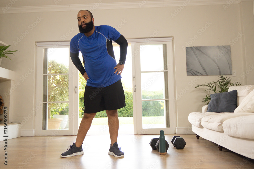 Fototapeta premium Adult African American man pausing in living room, wearing blue, hex dumbbells and green bottle