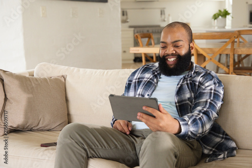 Adult African American man in plaid shirt sitting on sofa holding tablet, smiling at screen, phone