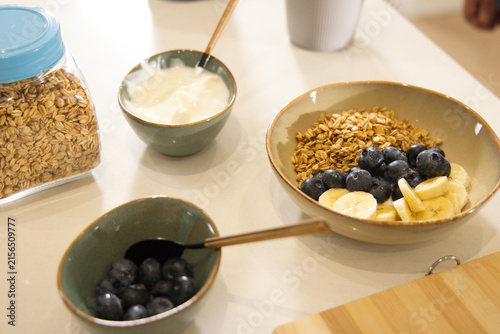 Granola bowl sitting on light counter with blueberries, banana, yogurt, oats jar and brass spoons