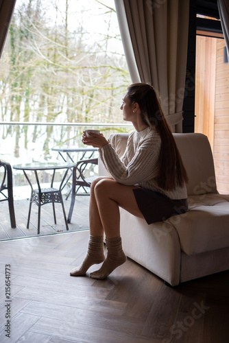 Woman with long hair sitting in an armchair by a large window in a country house, holding a mug of tea, wearing warm knitted beige sweater, brown skirt and cozy socks. Snowy winter forest outside.