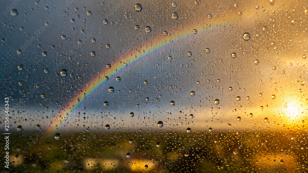 Fototapeta premium A vibrant rainbow appears through a rain-covered window at sunset, blending dark storm clouds with warm golden light.