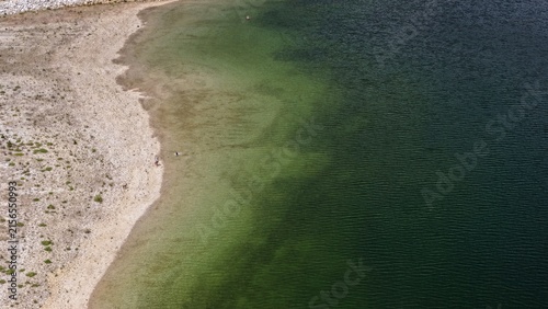 Aerial view of the shoreline of Twin Lakes with clear green water transitioning to deep blue and a pebbly beach with people walking along the water in Twin Lakes, Colorado, United States.