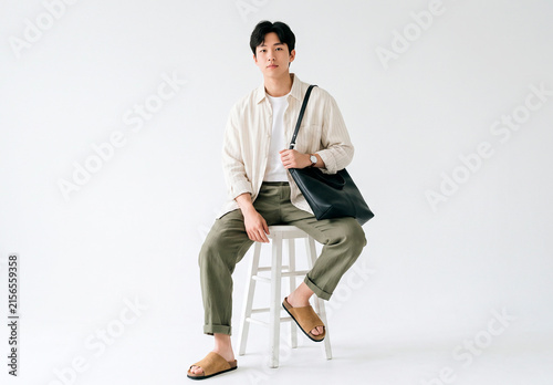 Handsome young Asian man in stylish casual outfit sitting on stool against white background. Fashionable male model posing with tote bag, wearing linen shirt and trousers. Minimalist studio portrait, 