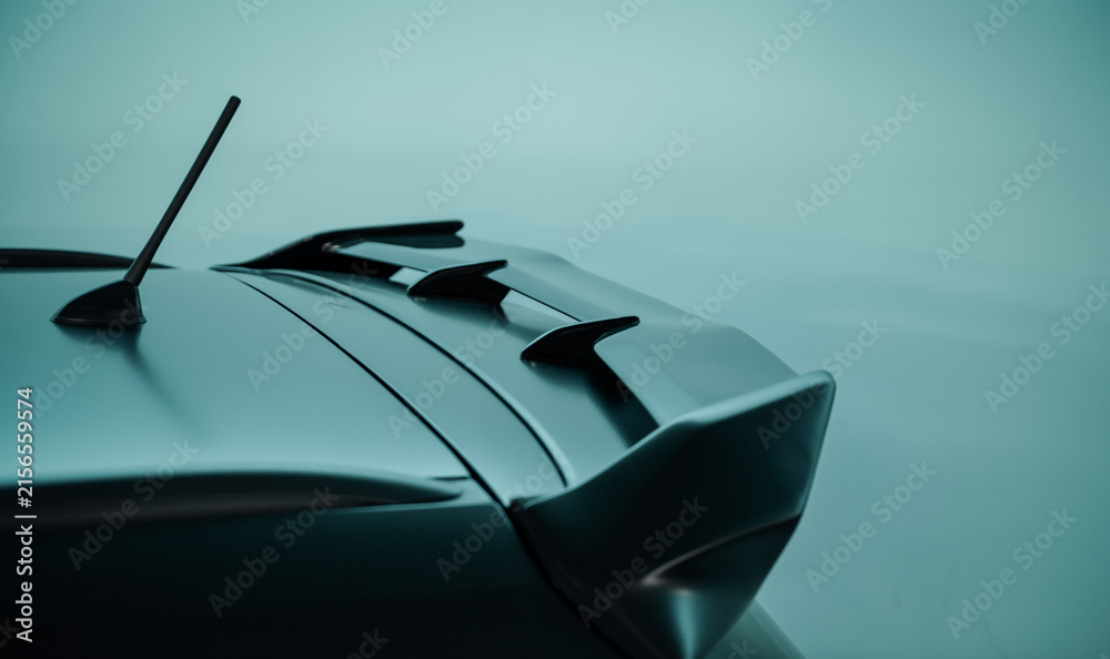 Fototapeta premium Rear spoiler on black car against bright teal background. Aerodynamic element on the top of car rear hatch for reducing lift.