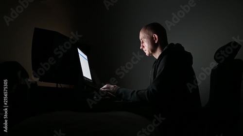 Hacker programming and typing on laptop computer at night his face illuminated by the screen in a dark clandestine room
