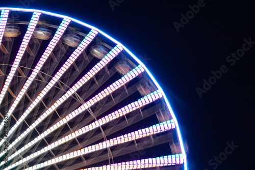 Long-exposure view of a brightly illuminated Ferris wheel against a dark night sky, with motion blur and colorful light trails creating a dynamic amusement park atmosphere.