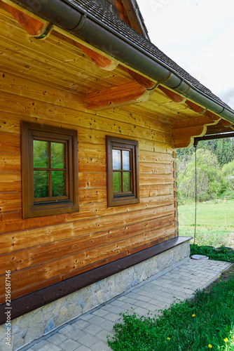 Rustic wooden cabin exterior with timber siding, traditional windows, stone foundation and a paved walkway overlooking a lush rural landscape.