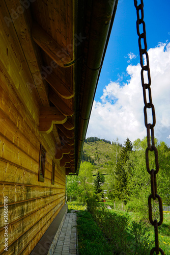Rustic timber cabin beside a lush mountain landscape, framed by a rain chain, wooden eaves, paved walkway, trees, and bright summer sky.