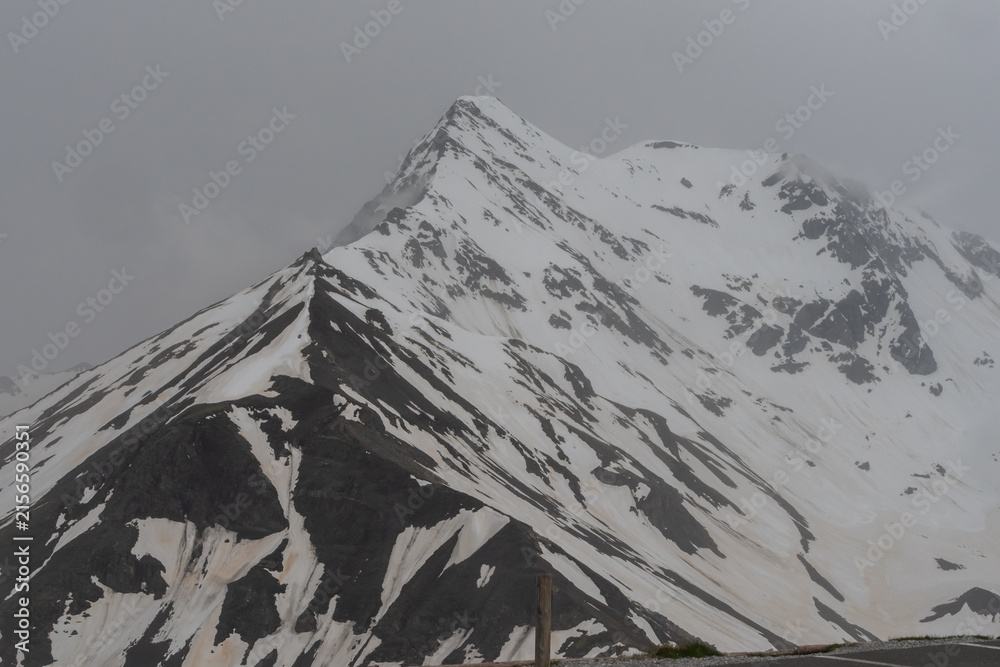 Obraz premium Picturesque Grossglockner High Alpine Road in Austria surrounded by rugged snowy peaks, green Alpine slopes and dramatic cloudy mountain scenery.