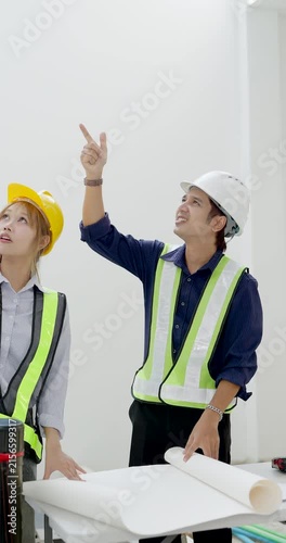 Asian female engineer and male contractor review large blueprint at table, pointing upward during construction progress meeting, safety planning, site document review, quality coordination indoors.