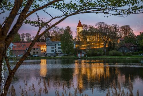 Scenic dawn over Talsi town in Latvia with church and lights reflecting in lake water, framed by tree branches
