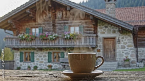 A cup of coffee sits on a wooden table outside a cabin in the mountains. The cabin has flowers on the balcony and a beautiful nature view. It is a peaceful moment in a rural setting