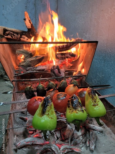 Fresh vegetables including bell peppers, tomatoes, eggplants, and onions grilling on skewers over open fire and hot coals in a metal barbecue.