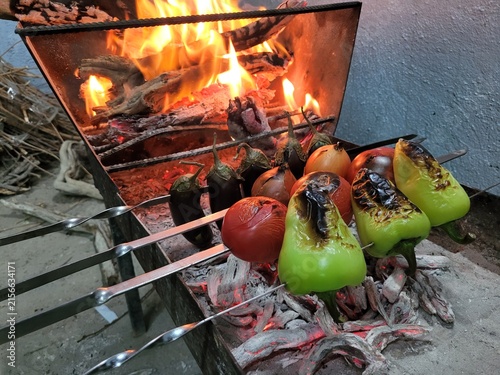 Fresh vegetables including bell peppers, tomatoes, eggplants, and onions grilling on skewers over open fire and hot coals in a metal barbecue.