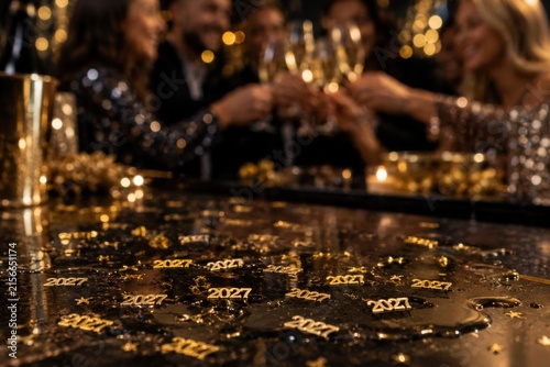 Golden 2027 confetti scattered across dark reflective table during lively group champagne toast party celebration event