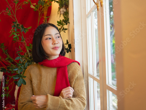 Konstfotografi Asian woman wearing brown sweater and red scarf looking out window in Christmas