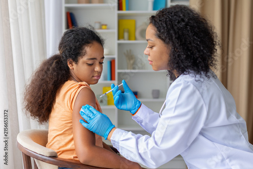 Fotografía Black female doctor giving young girl vaccination injection in the upper arm, il
